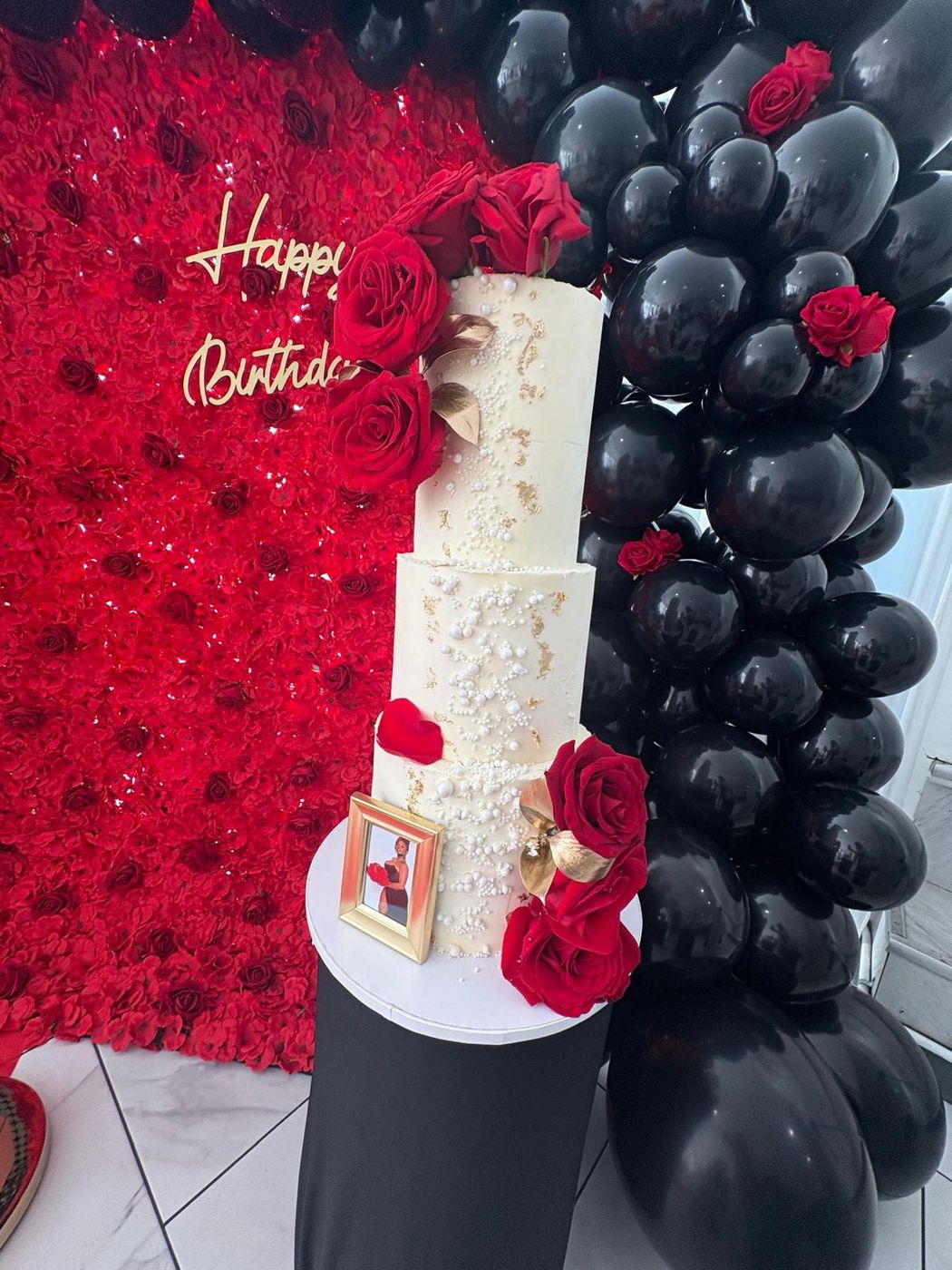 Tall white birthday cake with red roses and pearls in front of a red rose wall